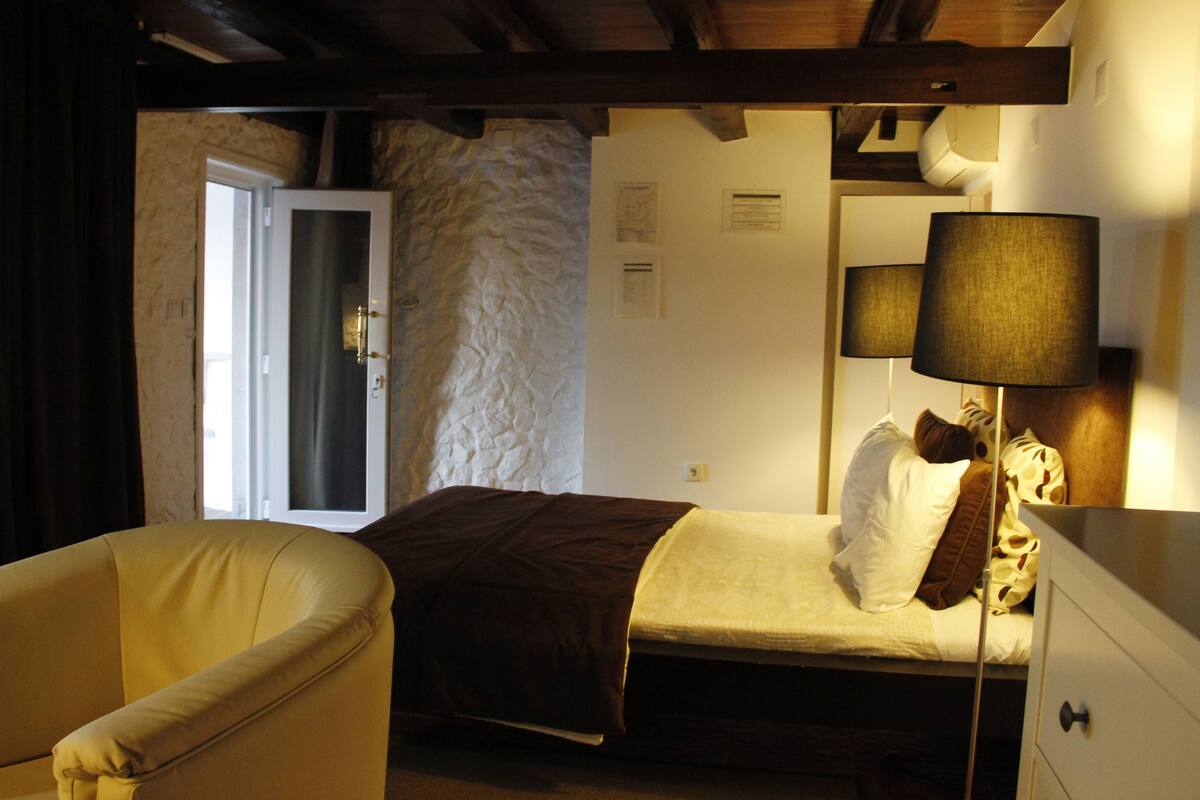 A bedroom with a bed, a chair, a lamp, and a door leading to another room.