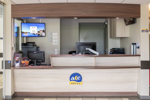 Un mostrador de recepción de hotel con un televisor de pantalla plana mostrando una vista panorámica, una mesa de recepción, una computadora y una caja fuerte.