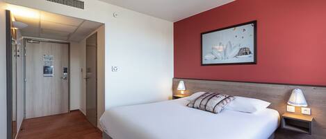 Habitación de hotel con cama, lámparas de noche, una pared de acento roja, un cuadro enmarcado y una puerta que da a otra habitación.