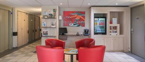 A lobby with red chairs, a reception desk, a painting, a refrigerator, and a coffee machine.