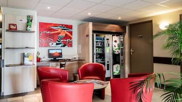 Un hall d’hôtel avec des fauteuils en cuir rouge, un comptoir d’accueil, un tableau, un distributeur automatique et un écran d’ordinateur.