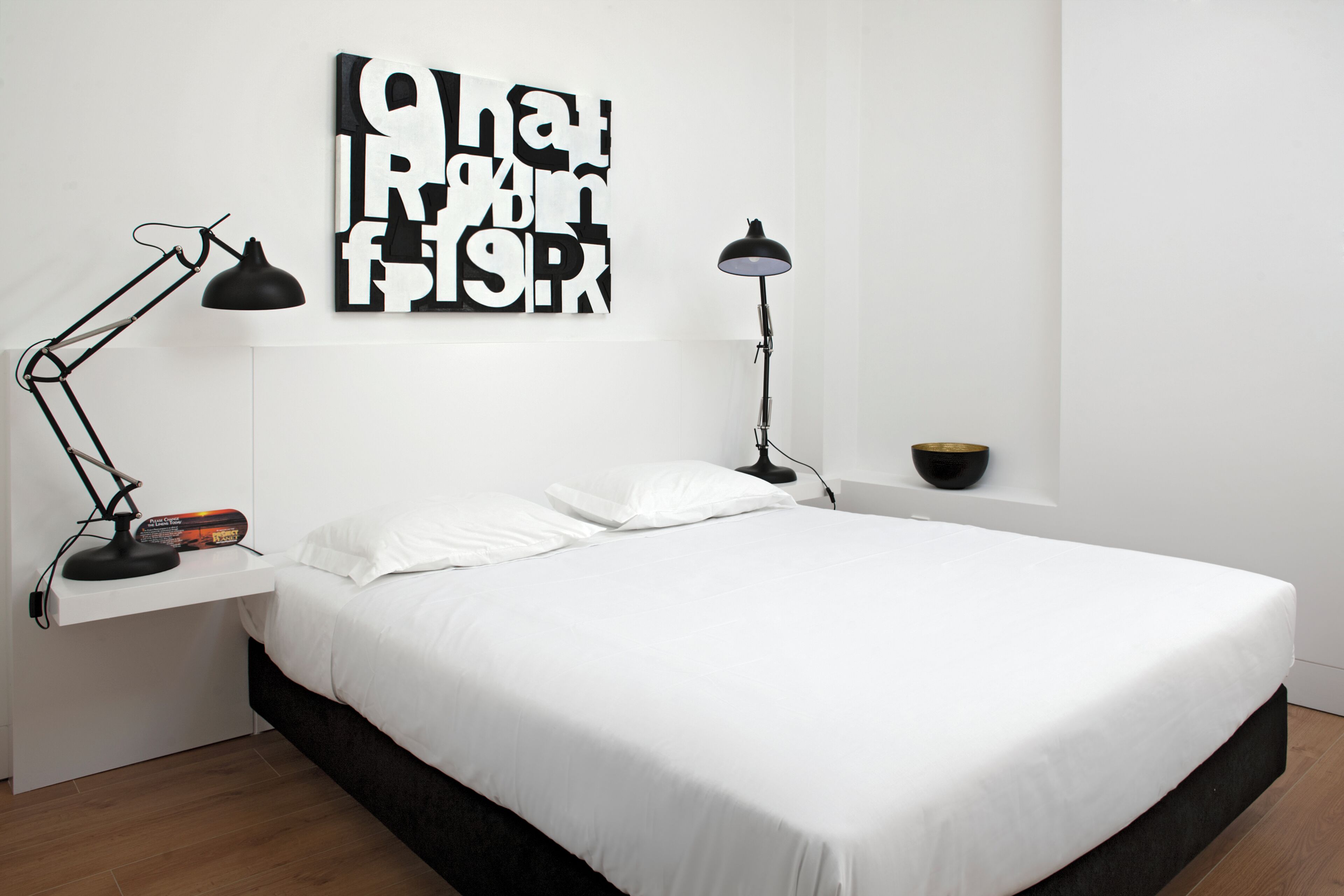 A bedroom with a bed, two bedside lamps, a wall-mounted artwork, and a small shelf with a lamp and a book.