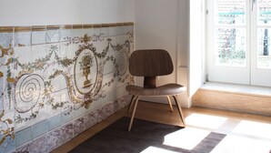 Una stanza con parete piastrellata, una sedia di legno, un tappeto e una finestra che si affaccia su un edificio.