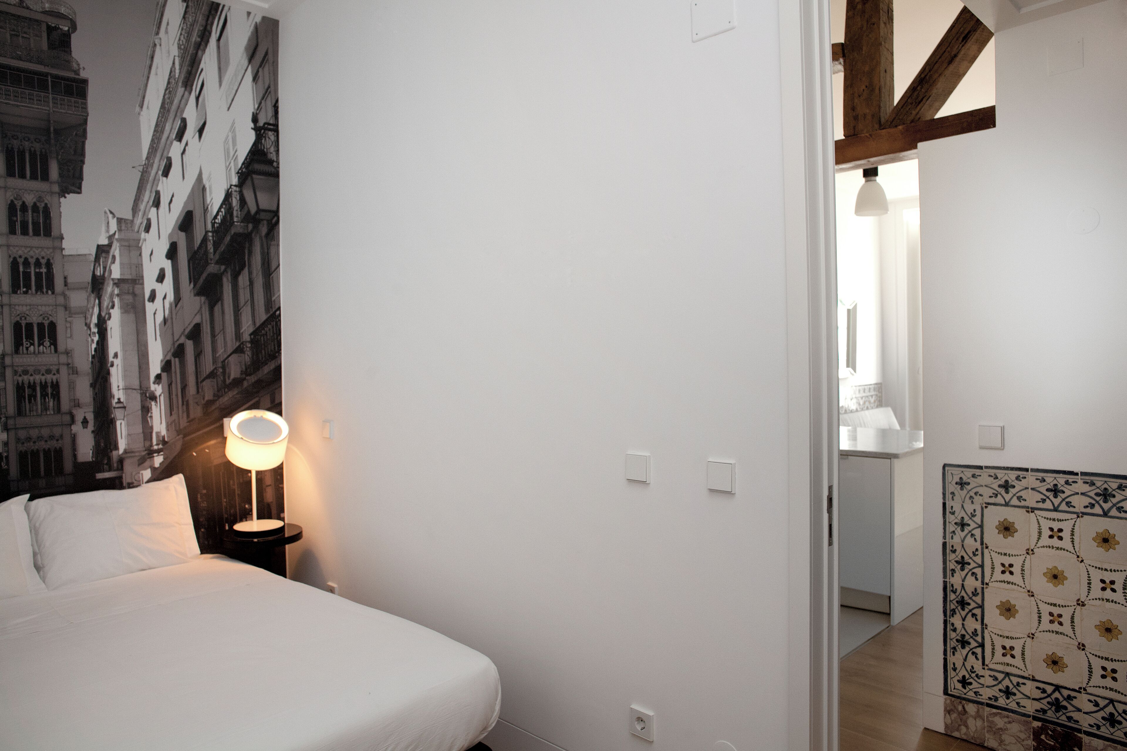 Un dormitorio con cama, una mesita de noche con lámpara, una pared decorada con azulejos y una puerta que da a otra habitación.
