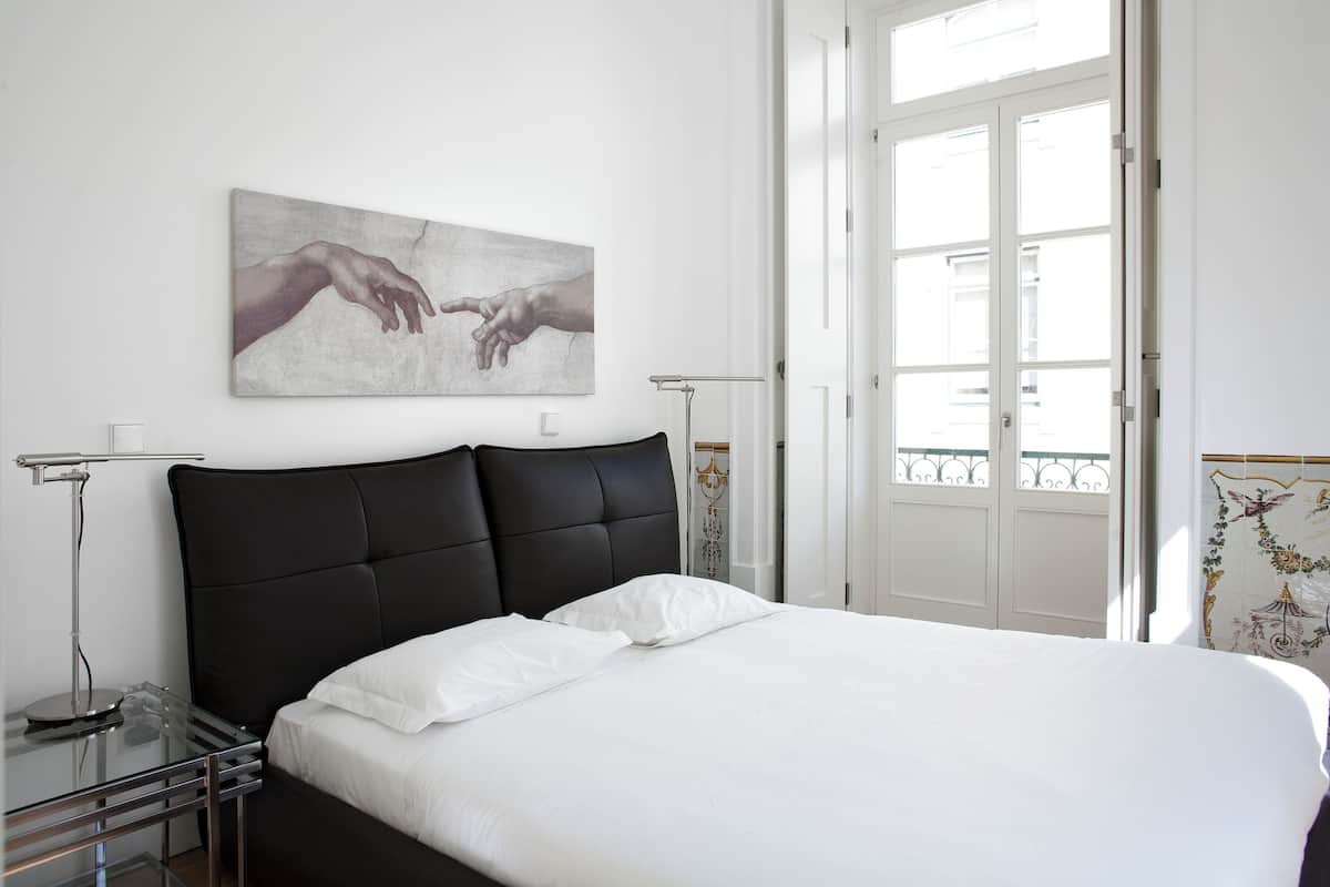 A bed with a black headboard, white bedding, a wall-mounted artwork of hands, a bedside table with a lamp, and a door with a balcony view.