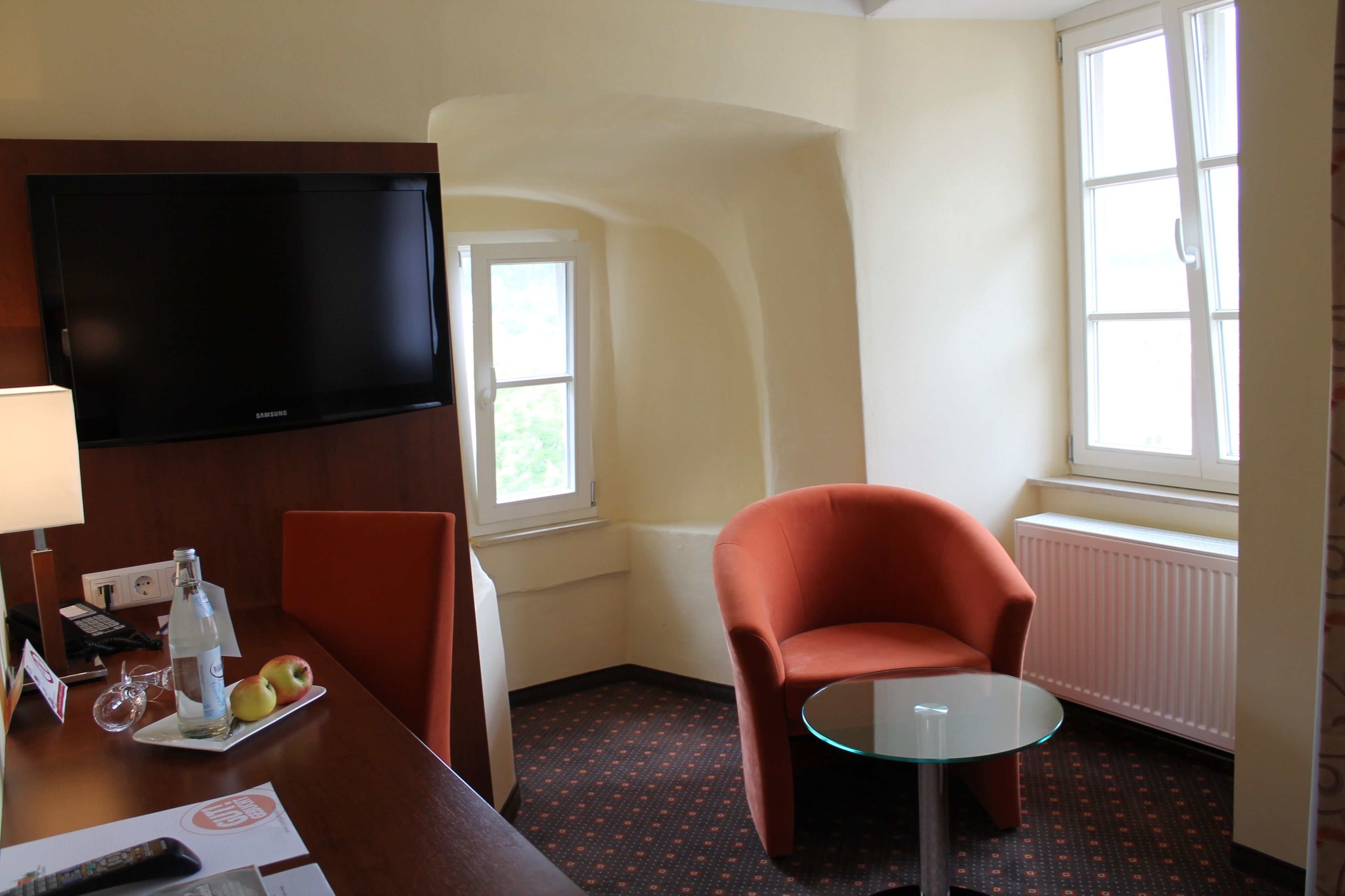 A hotel room with a flat-screen TV, a glass table, a red armchair, and a small dining table with apples and a bottle.