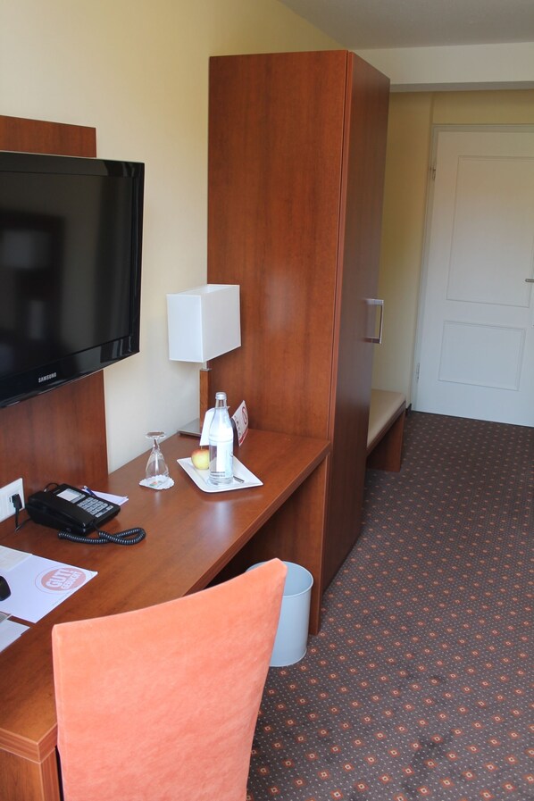 A hotel room with a wooden desk, a television, a lamp, a bottle of water, a glass, and a small table with a towel.