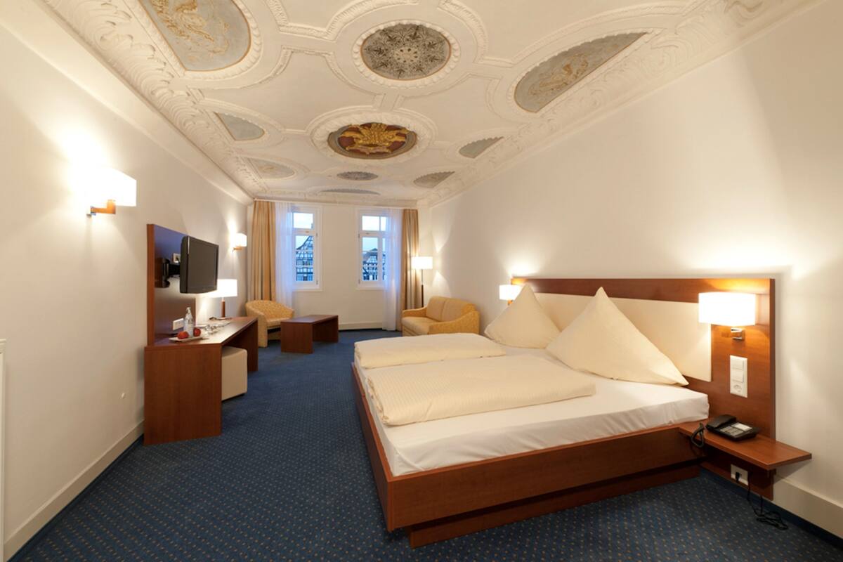 A hotel room with a large bed, a desk, a chair, a TV, and a view of buildings through the windows.
