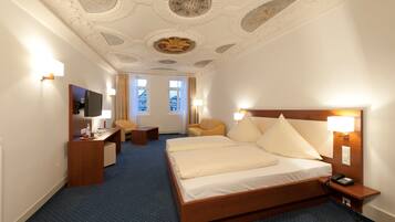 A hotel room with a large bed, a desk, a chair, a TV, and a view of buildings through the windows.