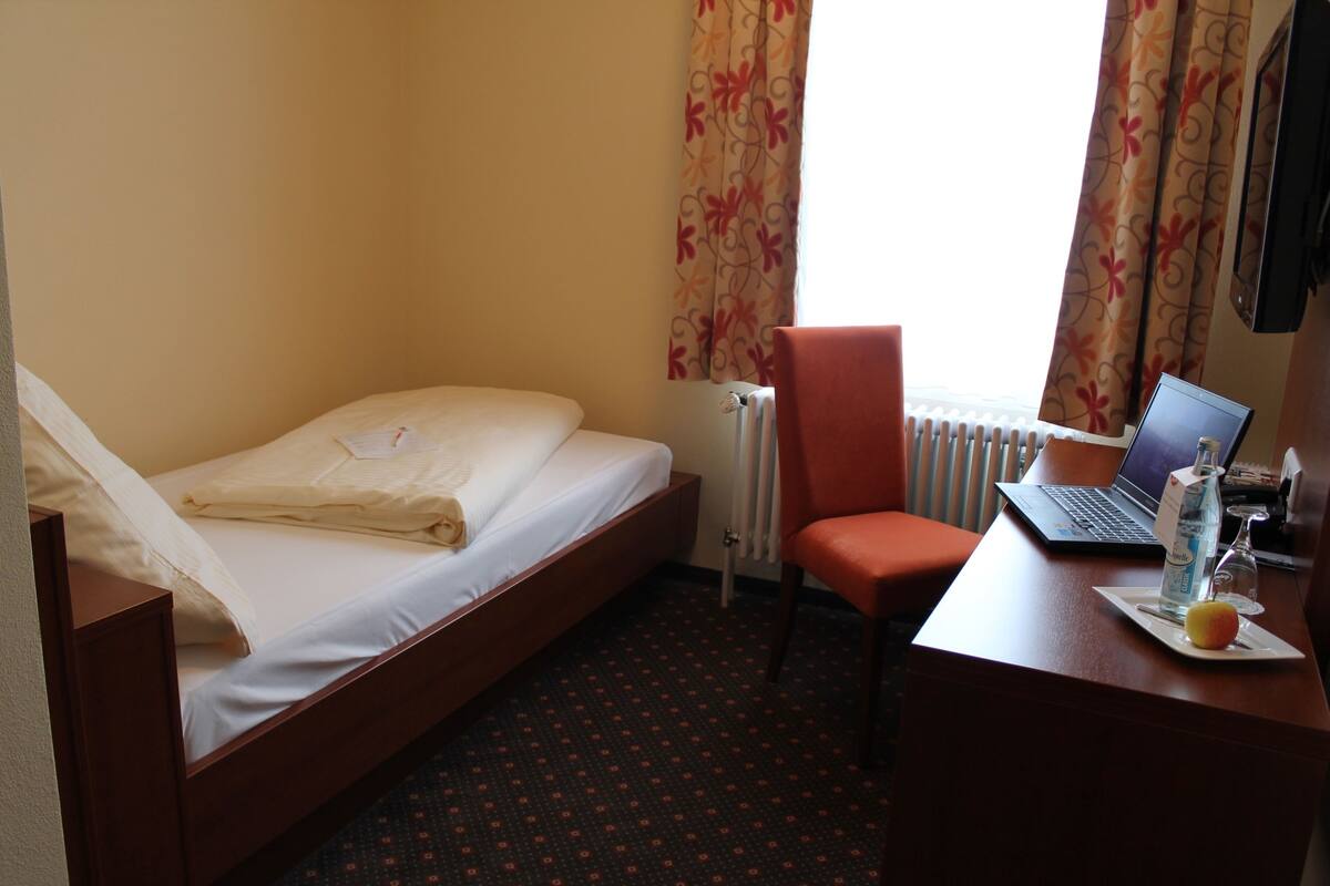 A hotel room with a single bed, a desk with a laptop, a chair, and a small table with a bottle and glass.