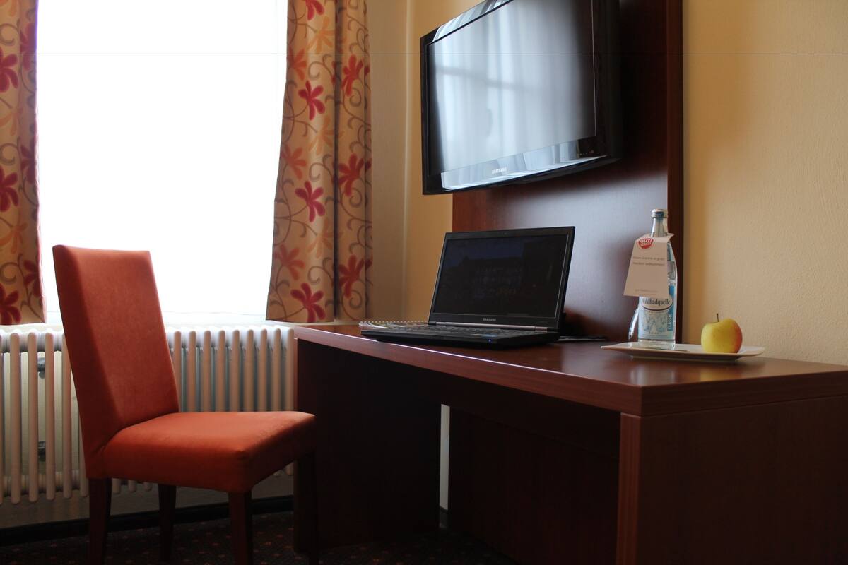 A hotel room with a wooden desk, a laptop, a bottle of water, a glass of water, a chair, a television, and a window with curtains.