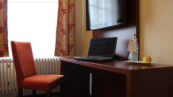 A hotel room with a wooden desk, a laptop, a bottle of water, a glass of water, a chair, a television, and a window with curtains.