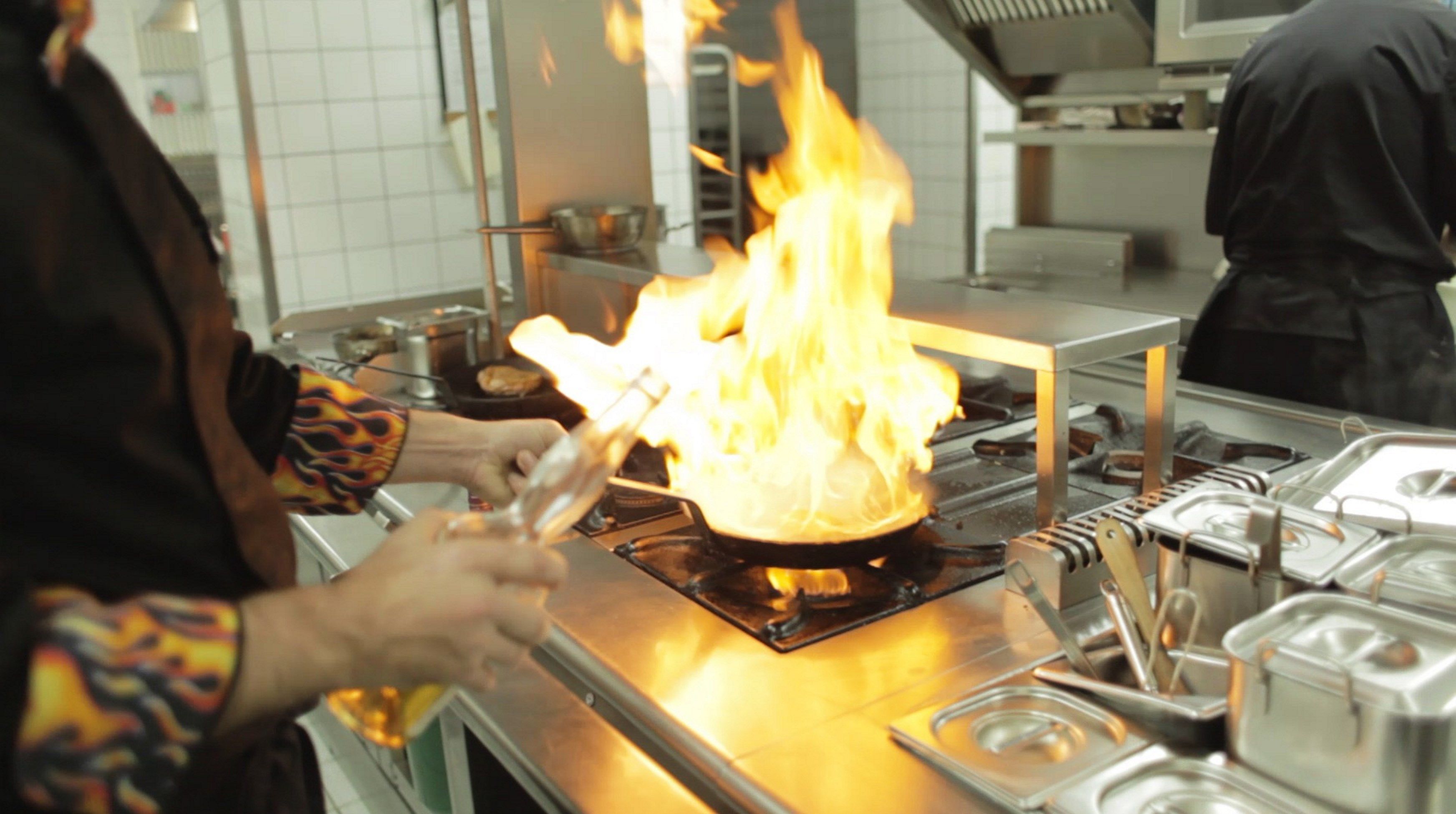 A kitchen with a stove on fire, a person handling a pan, and various kitchen equipment.