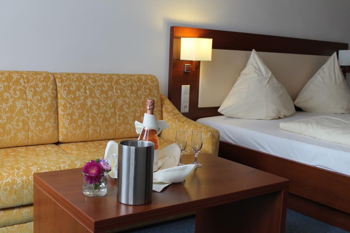 A hotel room with a wooden headboard, a bed with white linens, a yellow patterned sofa, a wooden nightstand with a glass vase of purple flowers, a bottle of water, and two wine glasses.