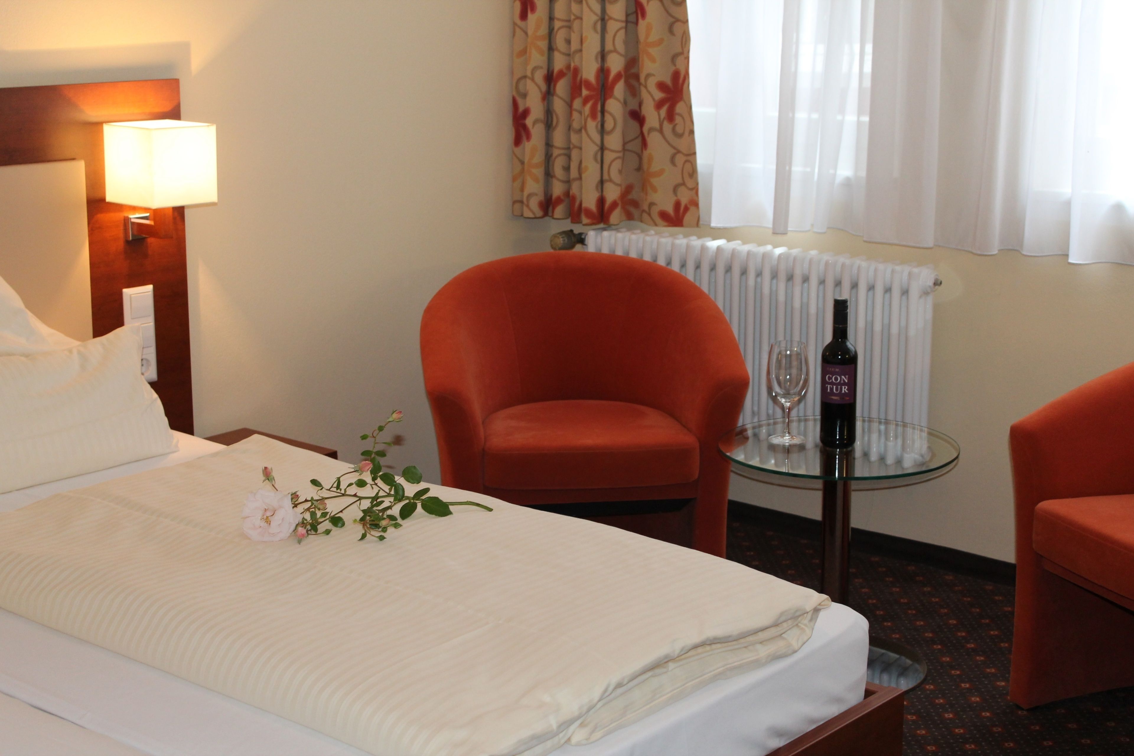 Ein Hotelzimmer mit einem Bett, zwei orangefarbenen Sesseln, einem kleinen Tisch mit einer Weinflasche, einer Lampe und einem Heizkörper.