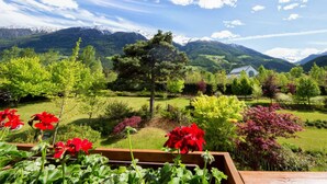 Un jardín con diversas plantas y flores, una terraza de madera y una casa al fondo con montañas.