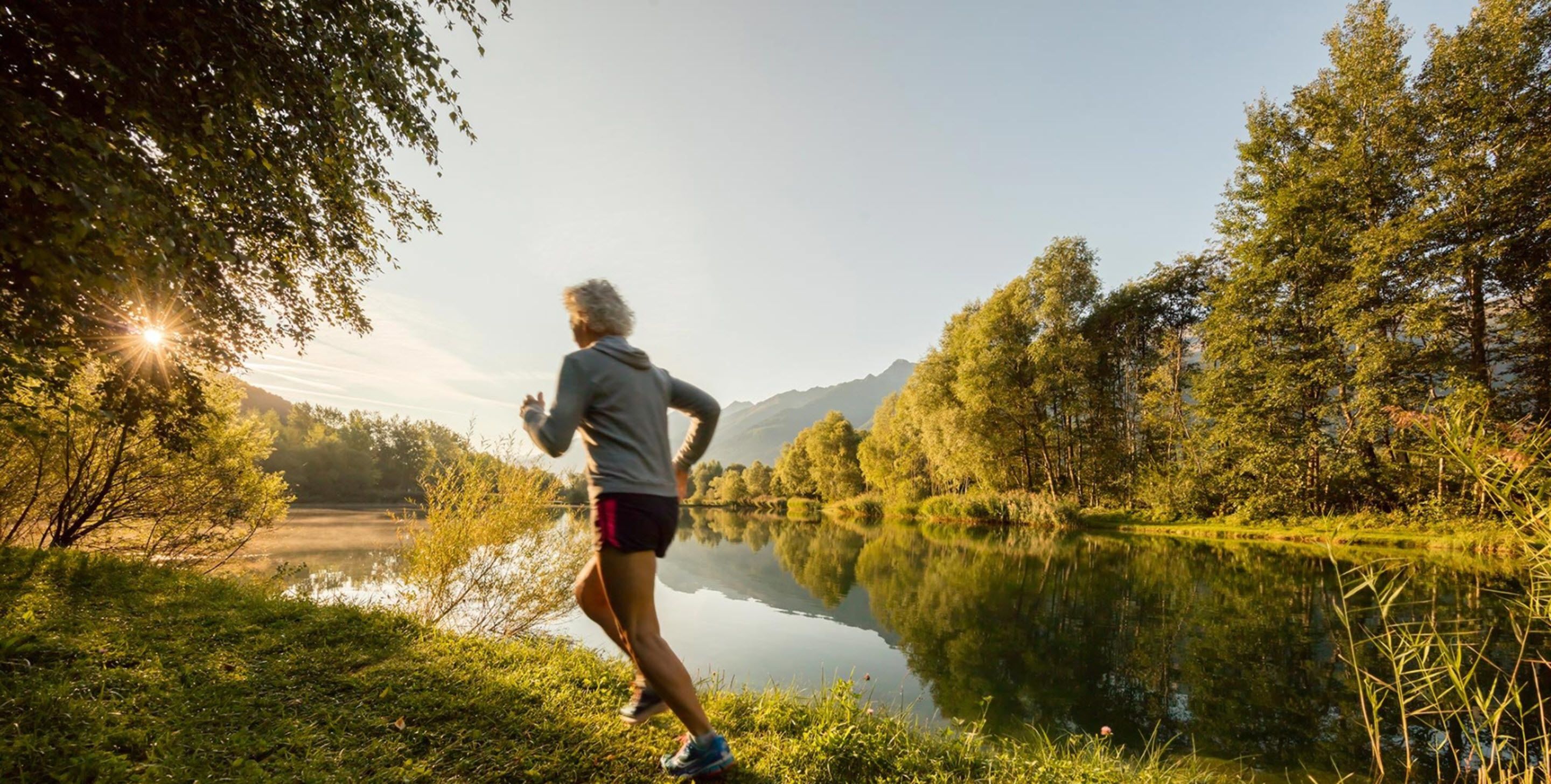 A person jogging along a lakeside path with trees and mountains in the background.