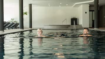 A modern indoor swimming pool with two people in the water, surrounded by a clean, minimalist interior with a staircase and potted plants.