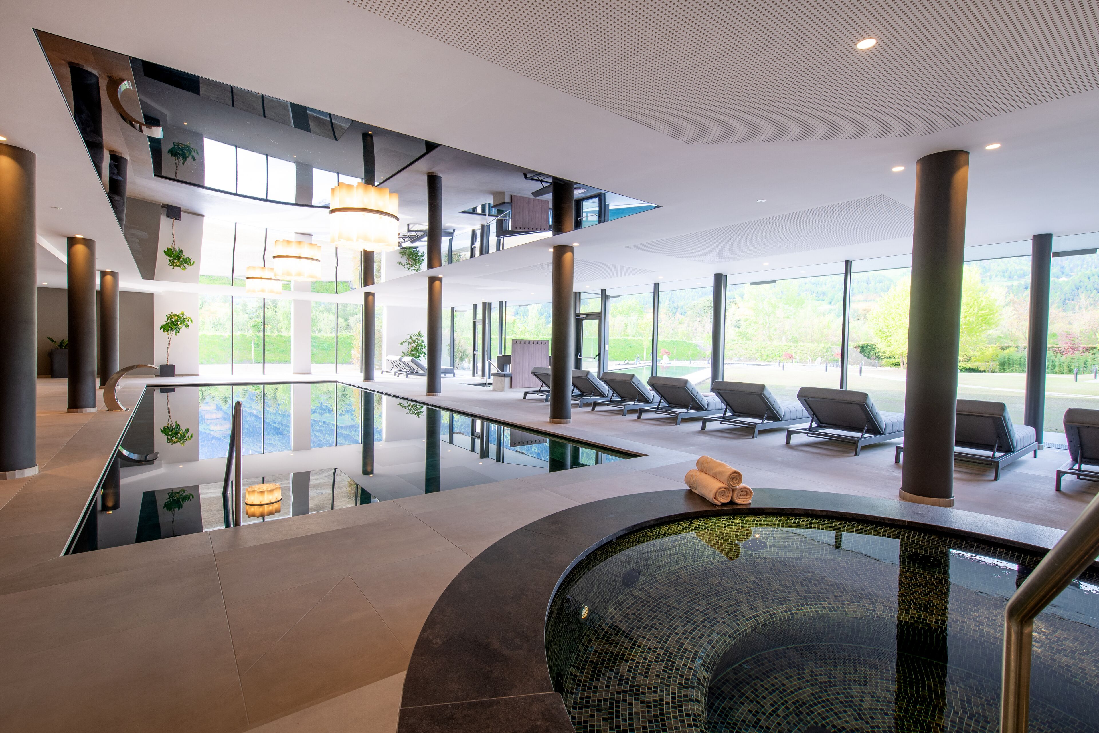 A modern indoor pool area with a glass barrier, lounge chairs, and a view of the outdoors.