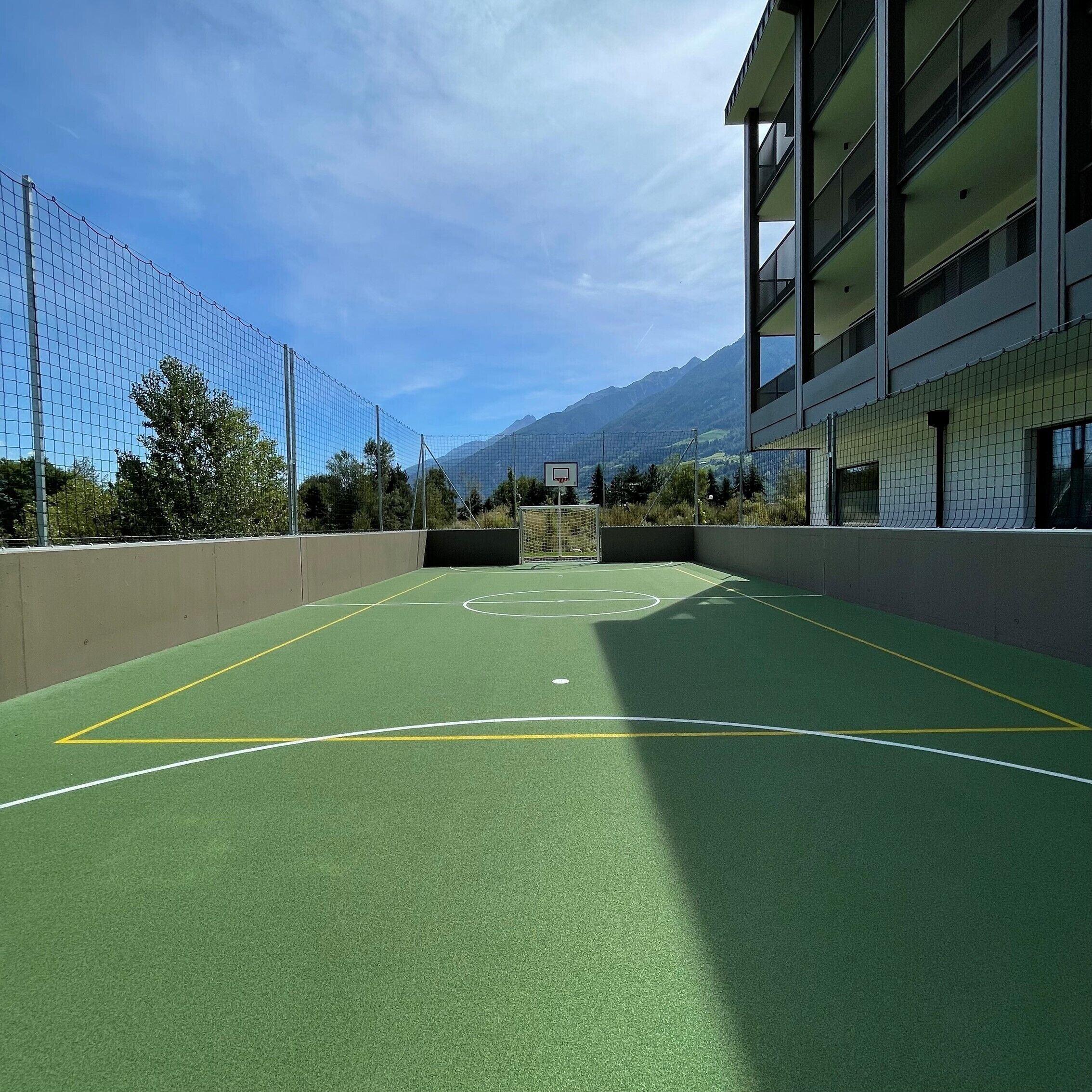 Una cancha de baloncesto al aire libre con valla de malla, aro de baloncesto y un edificio de varios pisos al fondo.