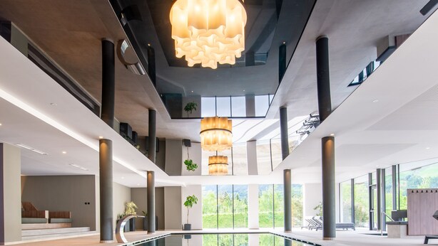 Un moderno vestíbulo de hotel con una piscina reflectante en el centro, grandes lámparas colgantes y un área de descanso con vistas a zonas verdes.