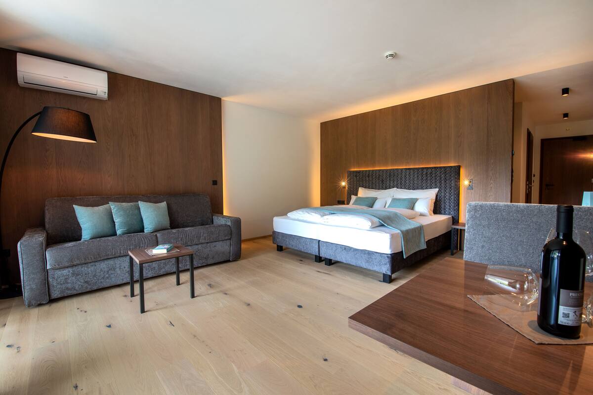 Une chambre d’hôtel moderne avec un panneau mural en bois, un canapé gris, un lit avec une tête de lit grise, une table basse en bois et une bouteille de vin posée sur la table.