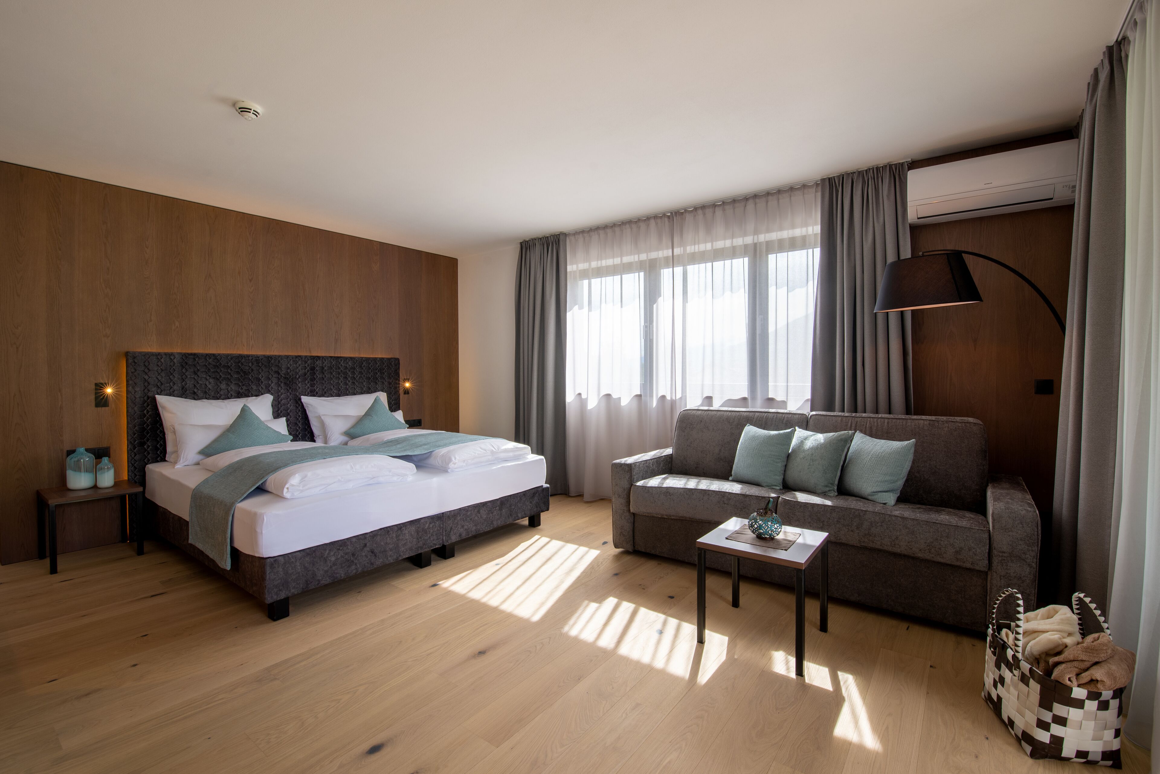 A modern hotel room with a large bed, a sofa, a small table, and a window with curtains.