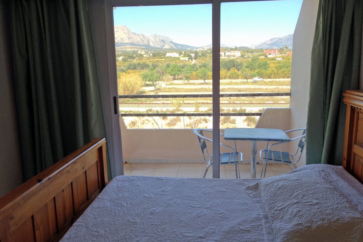 A bedroom with a bed, a balcony with a table and chairs, and a view of the countryside.