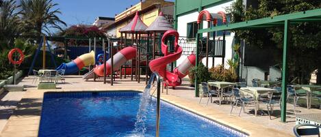 A swimming pool with a water slide, surrounded by lounge chairs and a playground with slides.