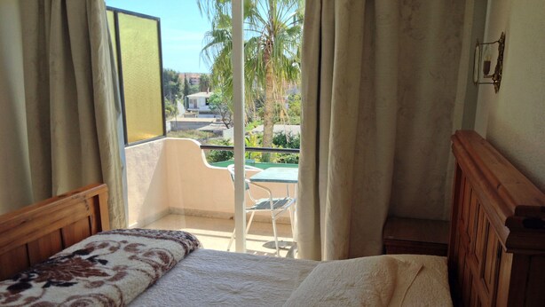 Un dormitorio con cama, cabecera de madera, ventana con cortinas y vistas a un balcón con silla y mesa.