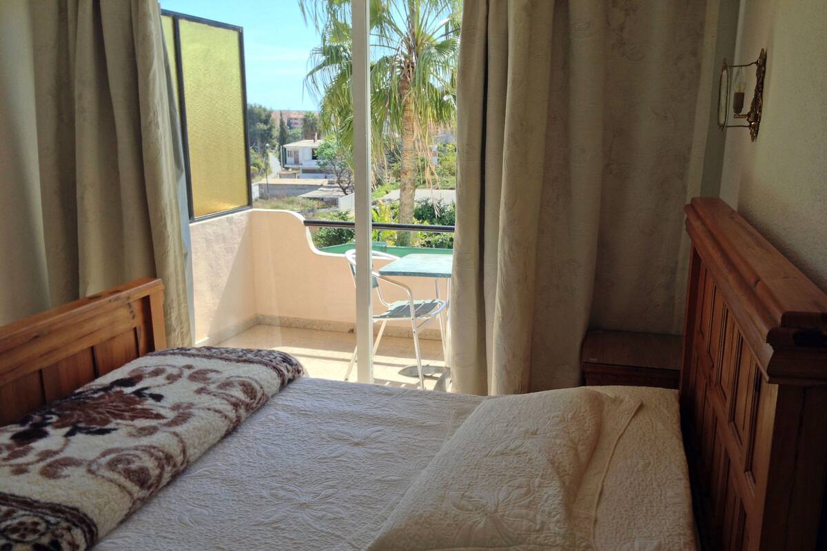 A bedroom with a bed, a wooden headboard, a window with curtains, and a view of a balcony with a chair and table.