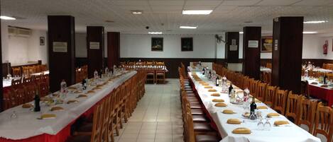 Un amplio salón de banquetes con largas mesas cubiertas de manteles blancos y caminos de mesa rojos, sillas de madera y piso de baldosas.