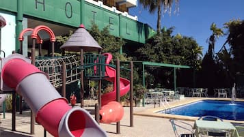A hotel with a children's play area including slides and climbing structures, a poolside seating area, and a clear blue sky.