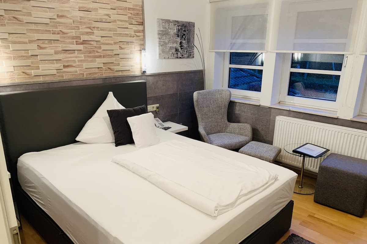 A modern hotel room with a large bed, a grey armchair, a small table, and a view of the outside through a window.