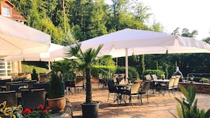 Un área de patio al aire libre con mesas y sillas bajo sombrillas blancas, rodeada de plantas en macetas y un muro de piedra.