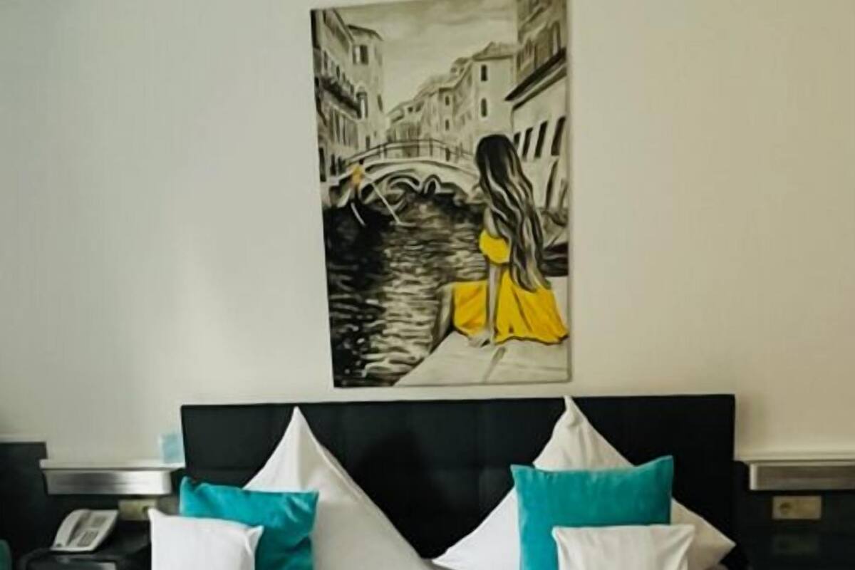 A hotel room with a bed, white bedding, teal pillows, and a painting of a woman in a yellow dress on a bridge.