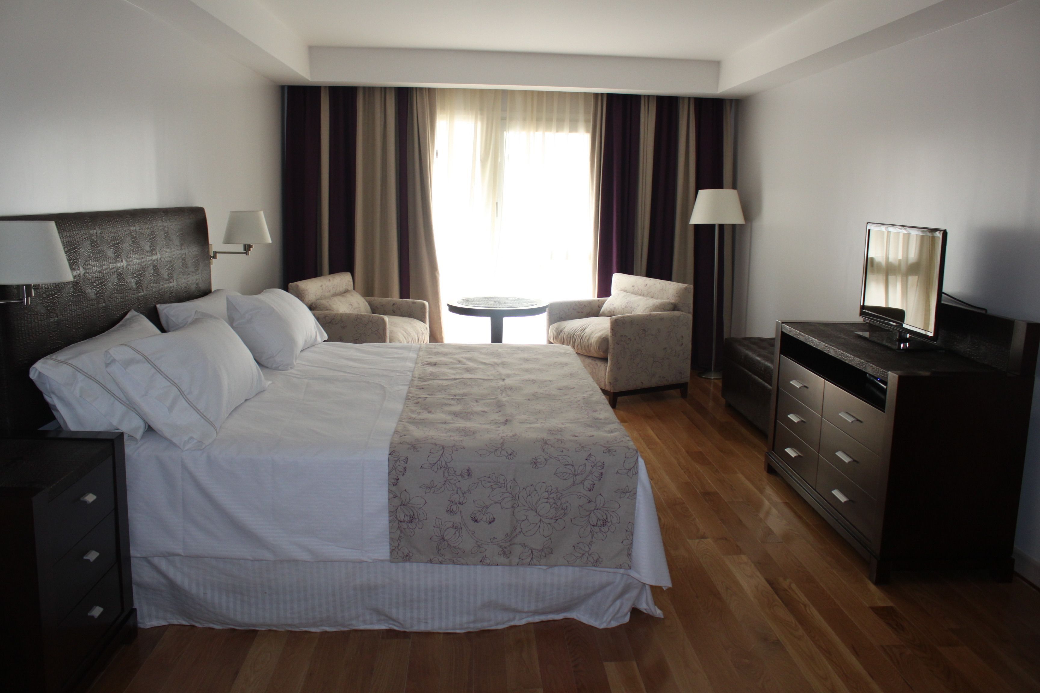 A hotel room with a large bed, two armchairs, a small round table, a TV, and a dresser.