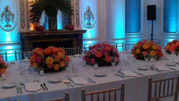 Una mesa de banquete dispuesta para un evento formal, con centros de mesa florales, manteles blancos y sillas de madera.