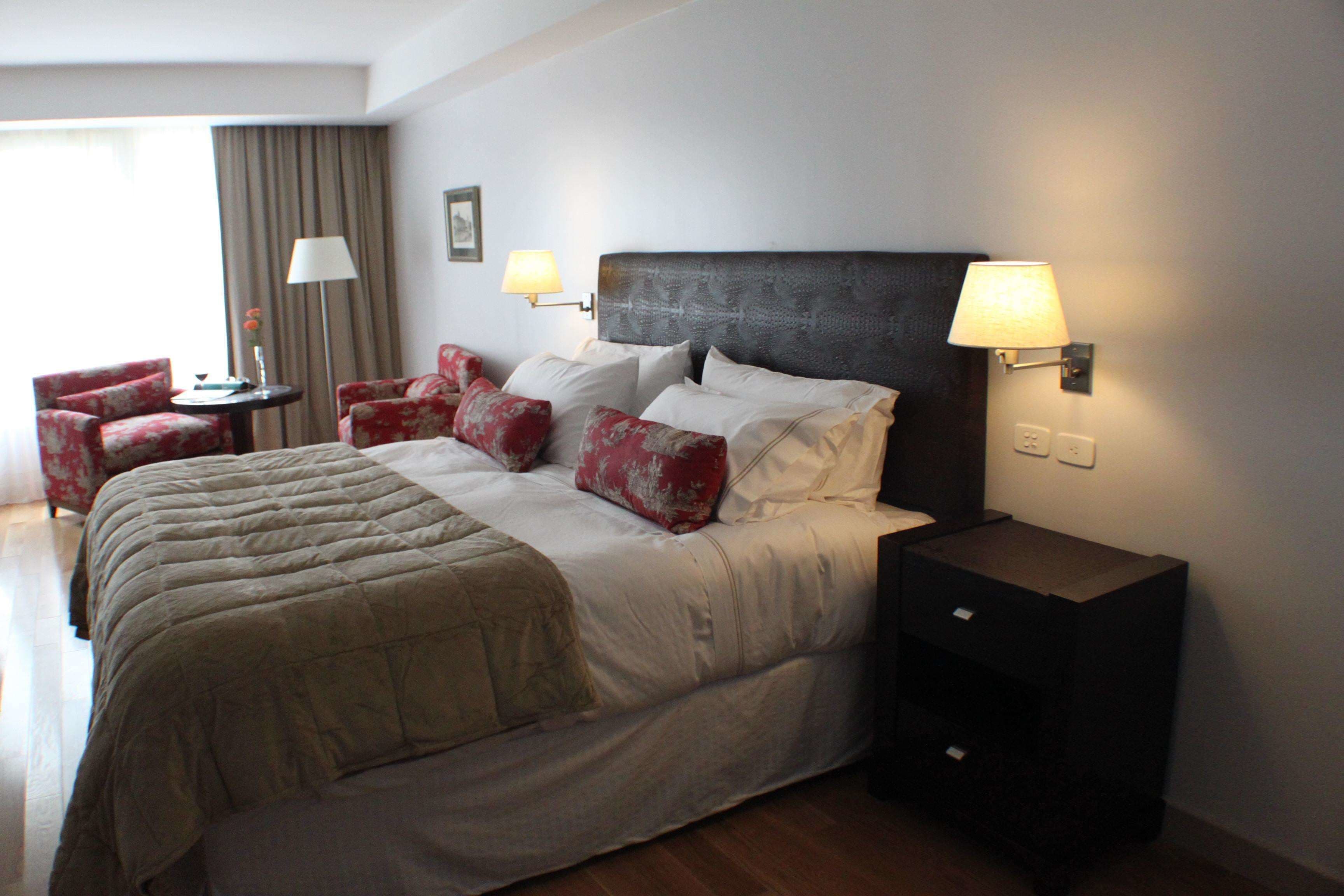 Habitación de hotel con una cama grande, dos lámparas de noche, una mesita pequeña y dos sillones rojos.