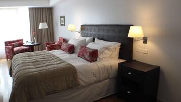 Habitación de hotel con una cama grande, dos lámparas de noche, una mesita pequeña y dos sillones rojos.