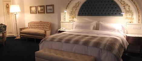 Una habitación de hotel con una cama grande, una zona de estar con una silla, una mesita de noche con lámpara y cuadros enmarcados en la pared.