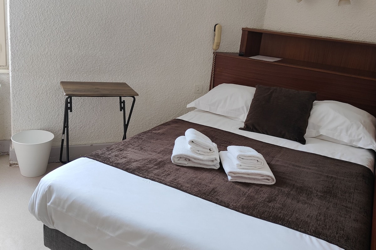 A hotel room with a bed, a wooden headboard, a painting of a landscape, a small table, and a white bucket.