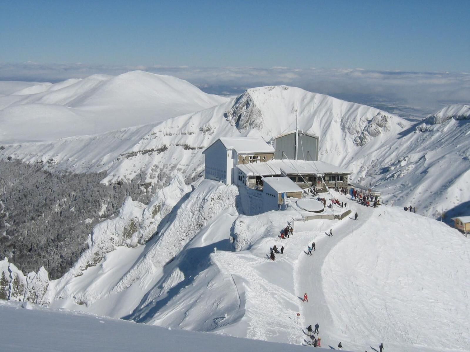 A snow-covered mountain with a lodge featuring a large building and a smaller structure, surrounded by snow-covered slopes and a clear sky.