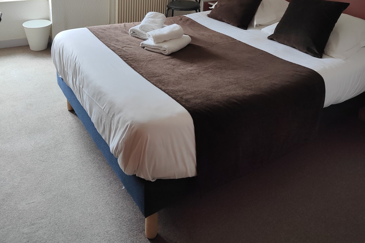 A neatly made bed with a brown and white duvet, a chair, and a radiator in a room with a sloped ceiling and a window.