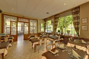 A dining area with tables set for service, large windows with curtains, and a view of the outdoors.