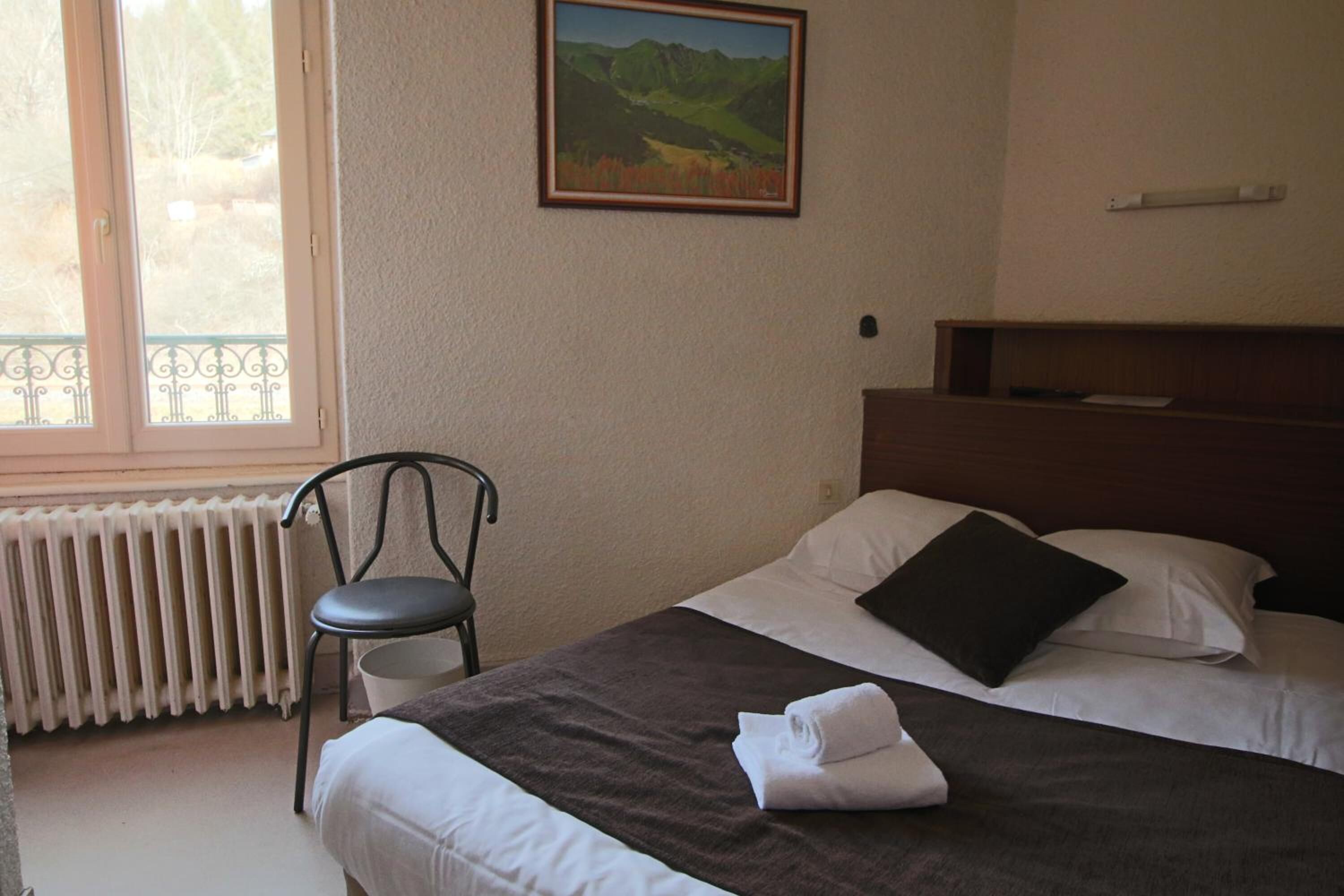 Hotelkamer met een bed, een stoel, een radiator, een schilderij aan de muur en een raam met uitzicht op groen.