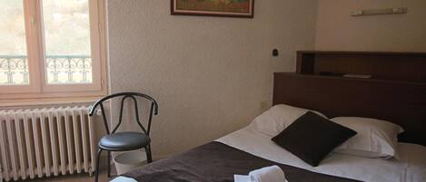 A hotel room with a bed, a chair, a radiator, a painting on the wall, and a window with a view of greenery.