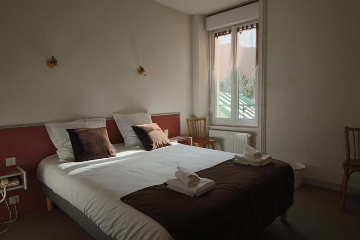 A neatly made bed with white and brown linens, a wooden nightstand with a telephone, and a window with a view of greenery.