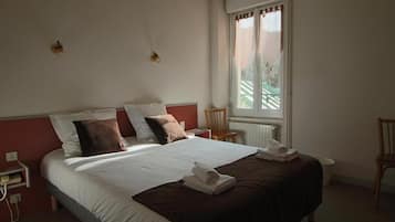 Una cama bien tendida con ropa de cama blanca y marrón, una mesita de noche de madera con un teléfono y una ventana con vistas a zonas verdes.