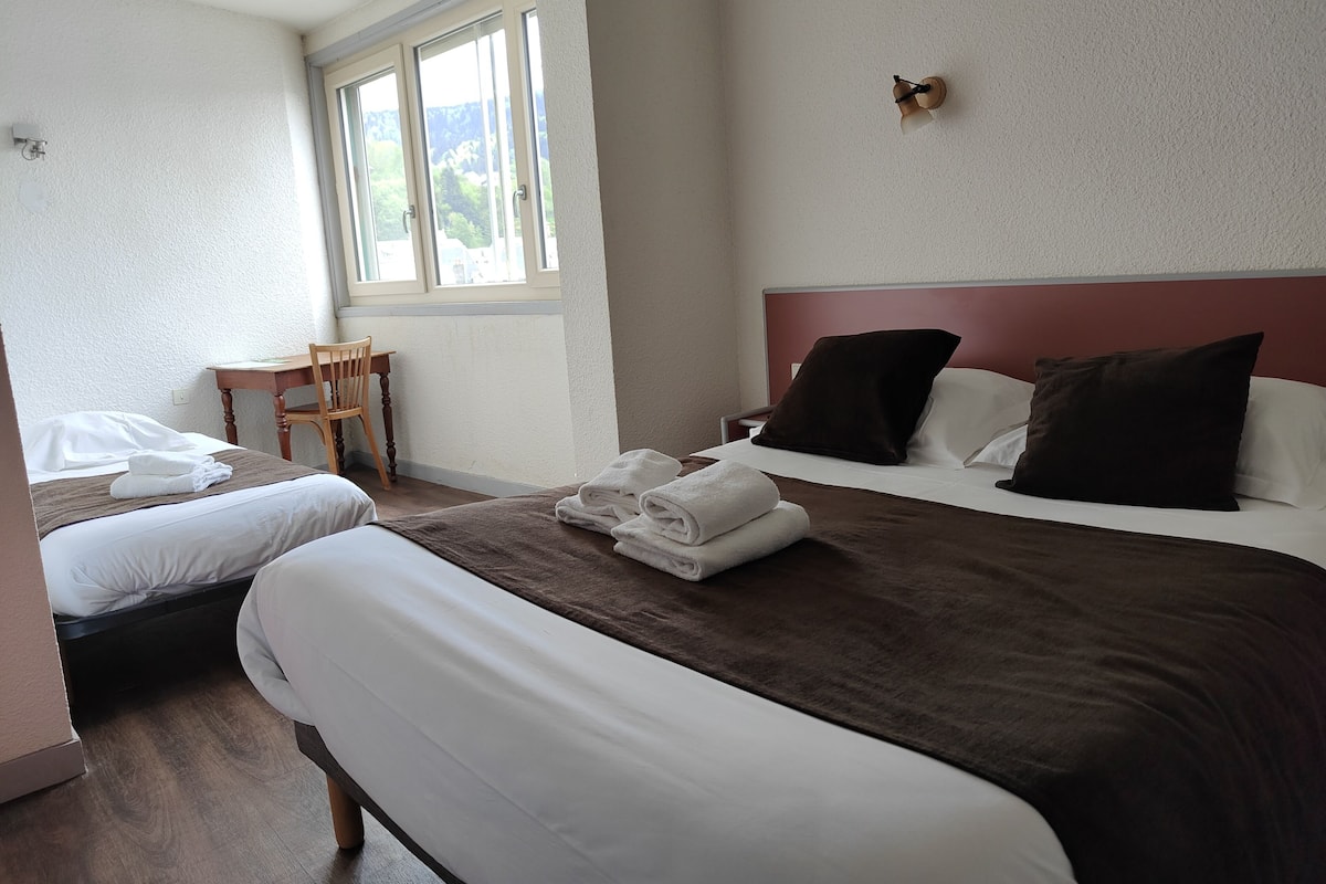 A hotel room with two beds, a wooden chair, a table, and a window with a view of greenery.